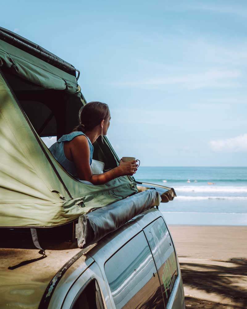 Woman lies in a rooftop tent drinking coffee